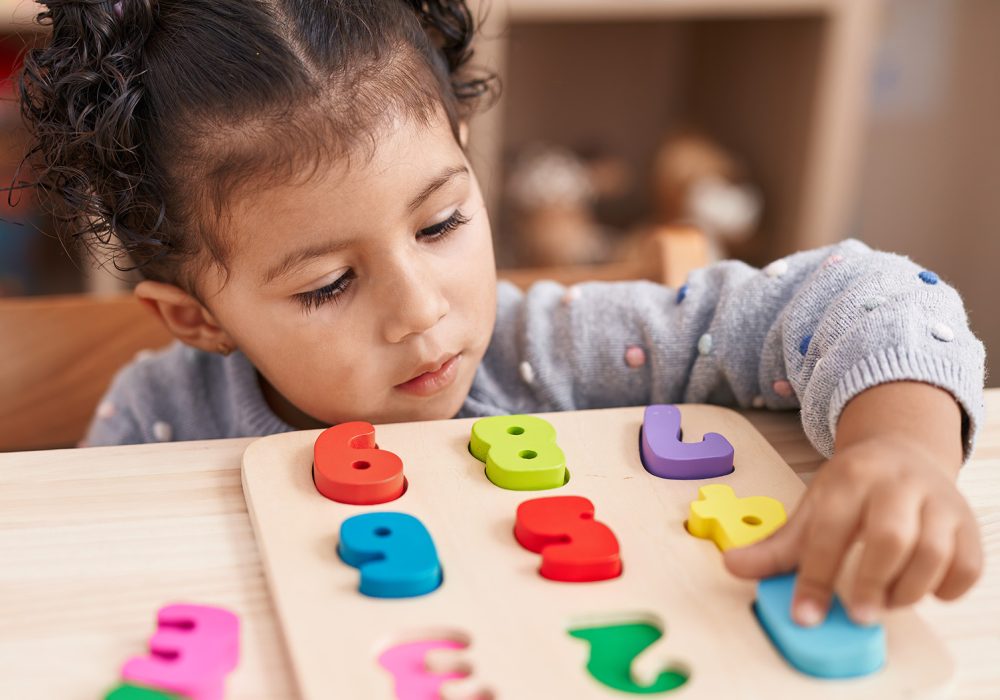 Adorable hispanic girl playing with maths puzzle game sitting on table at kindergarten